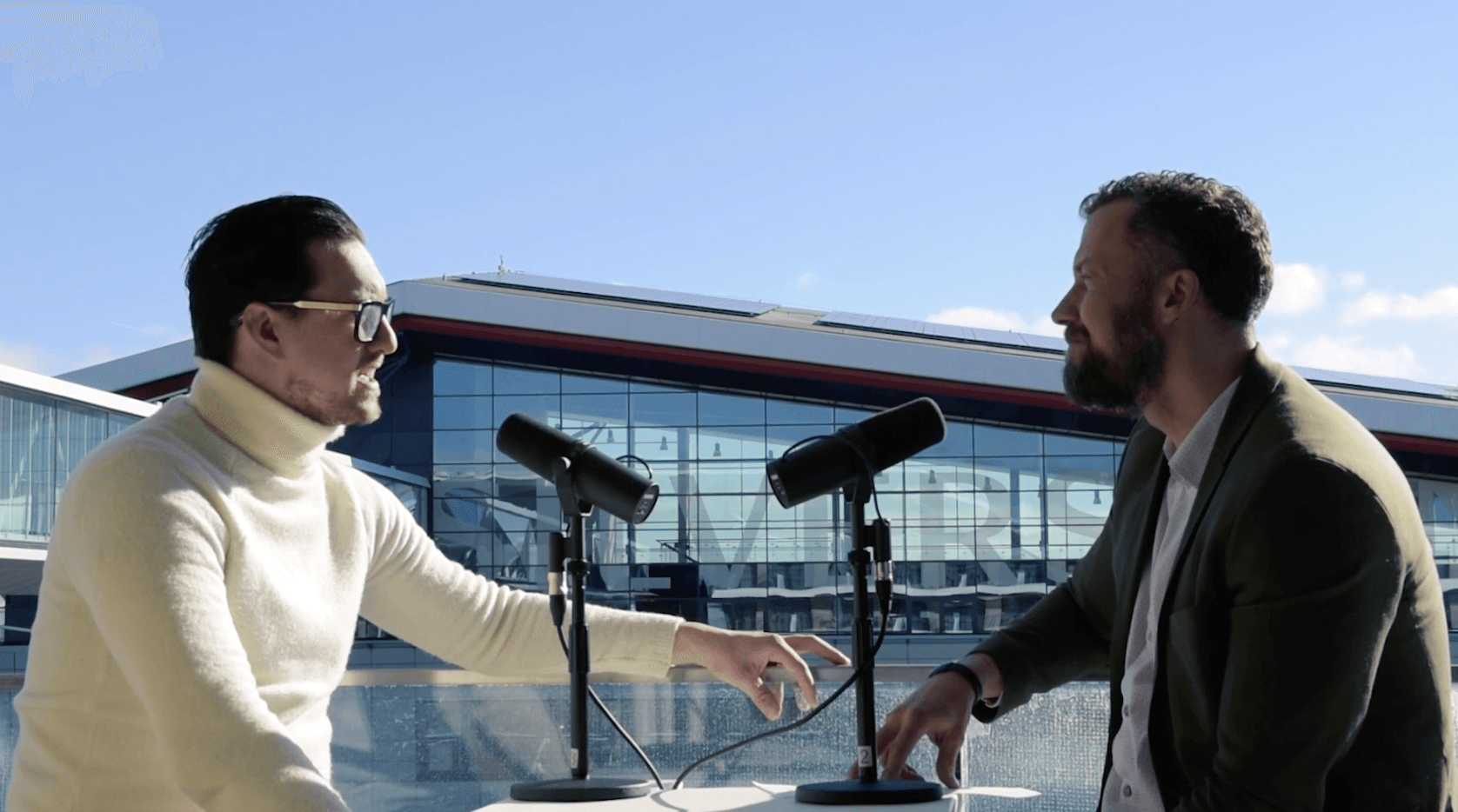 Two men in conversation across a desk with podcast microphones